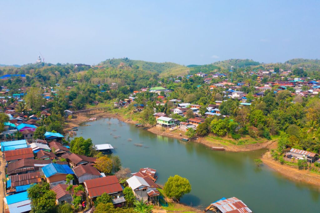 Aerial Top View Of Residential Local Houses In Mon Village, Nature Trees With Lake Or River, Kanchanaburi, Thailand In Urban City Town In Asia, Buildings.